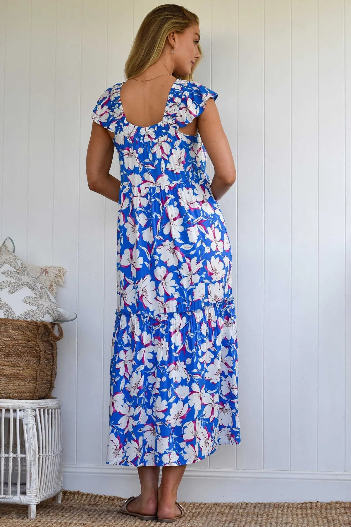 A woman standing from the back looking to the side, wearing a blue midi dress with a white and pink floral pattern, cap sleeves, and an open-back design.