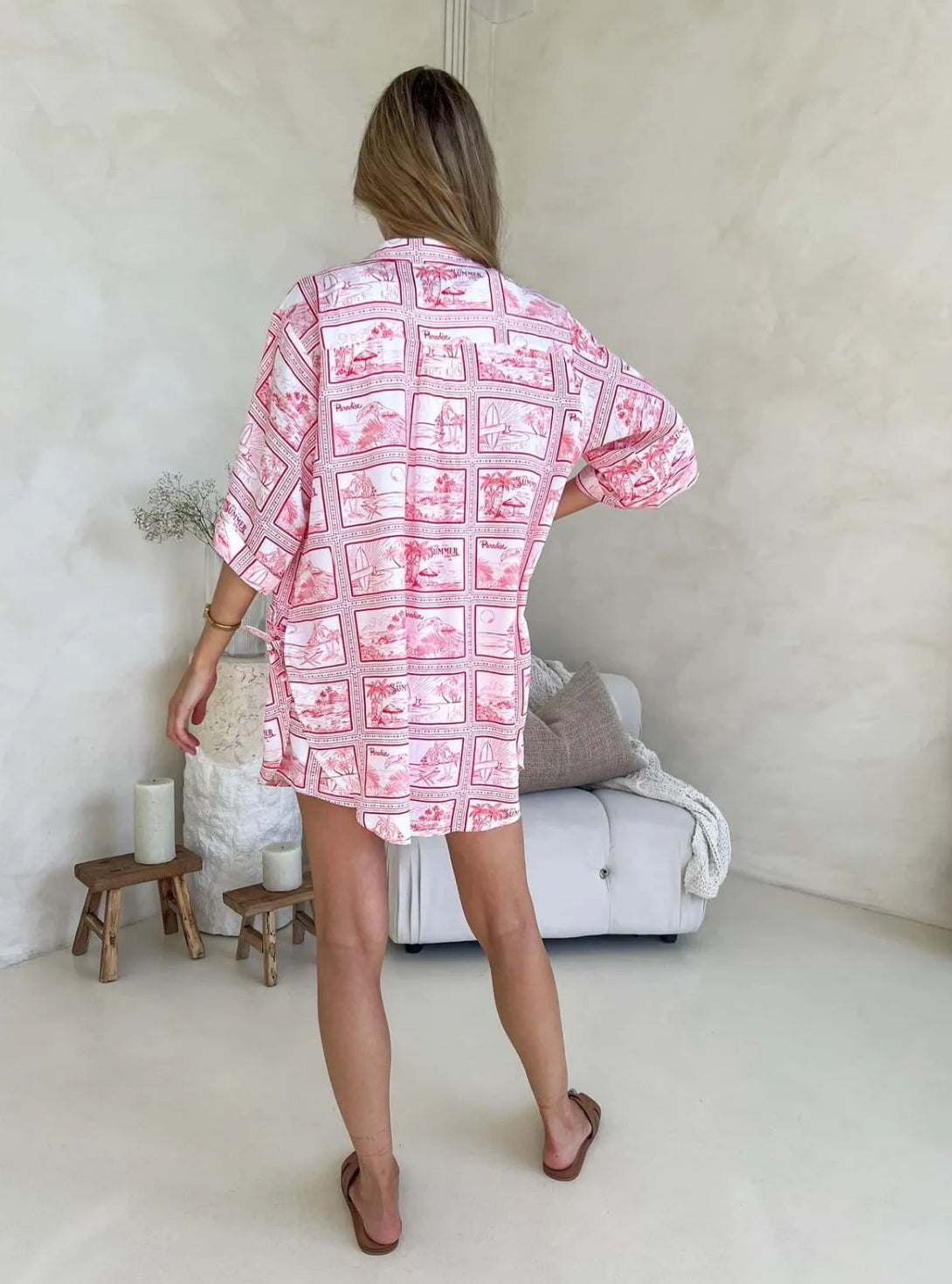 Back view of a woman wearing a red and white postcard print two-piece set, consisting of a button-up shirt and drawstring shorts, standing in a room with a light-colored wall and a chair in the background.