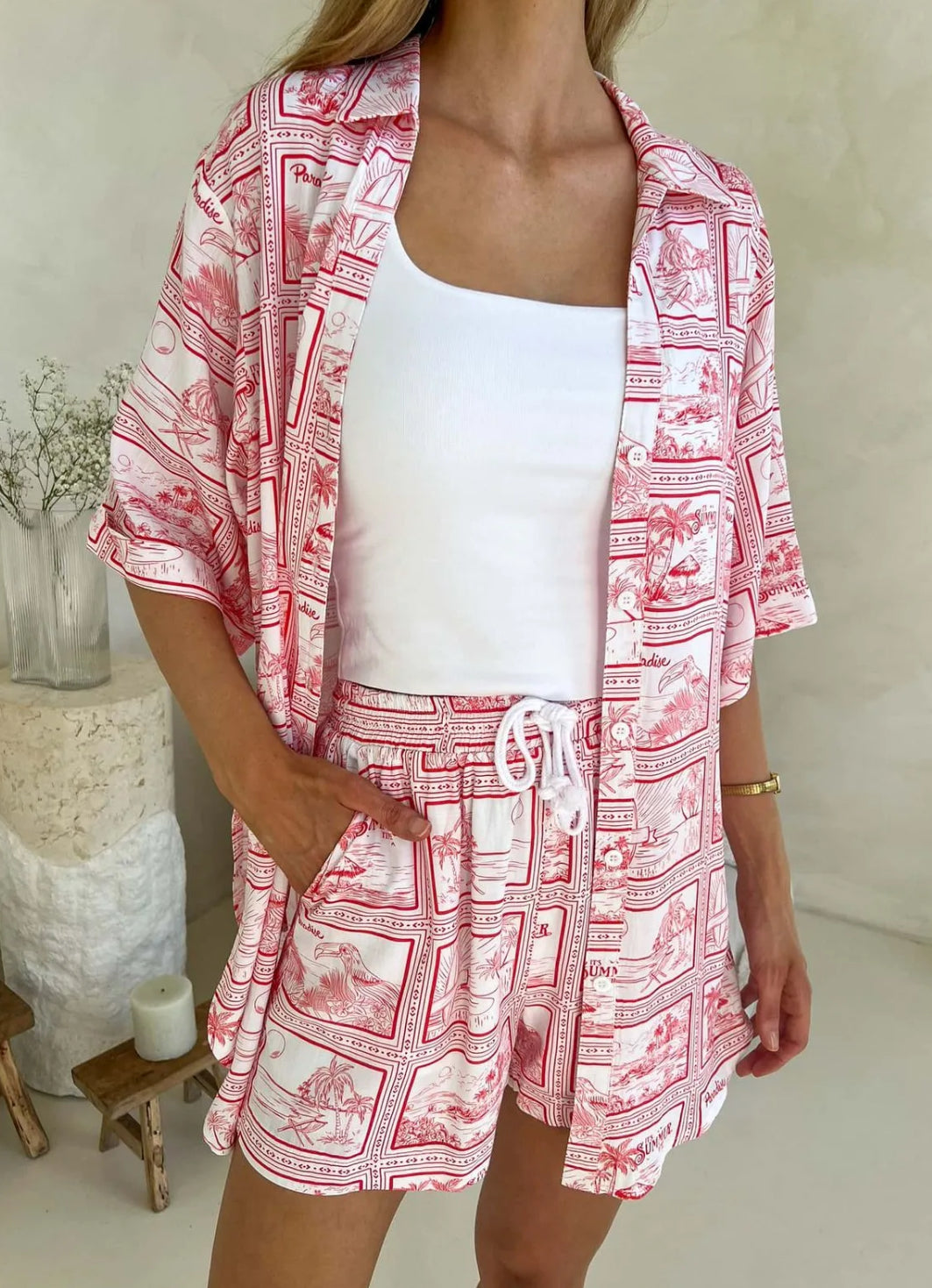A woman wearing a red and white postcard print two-piece set, consisting of a button-up shirt and drawstring shorts, standing in a room with a light-colored wall and a chair in the background.