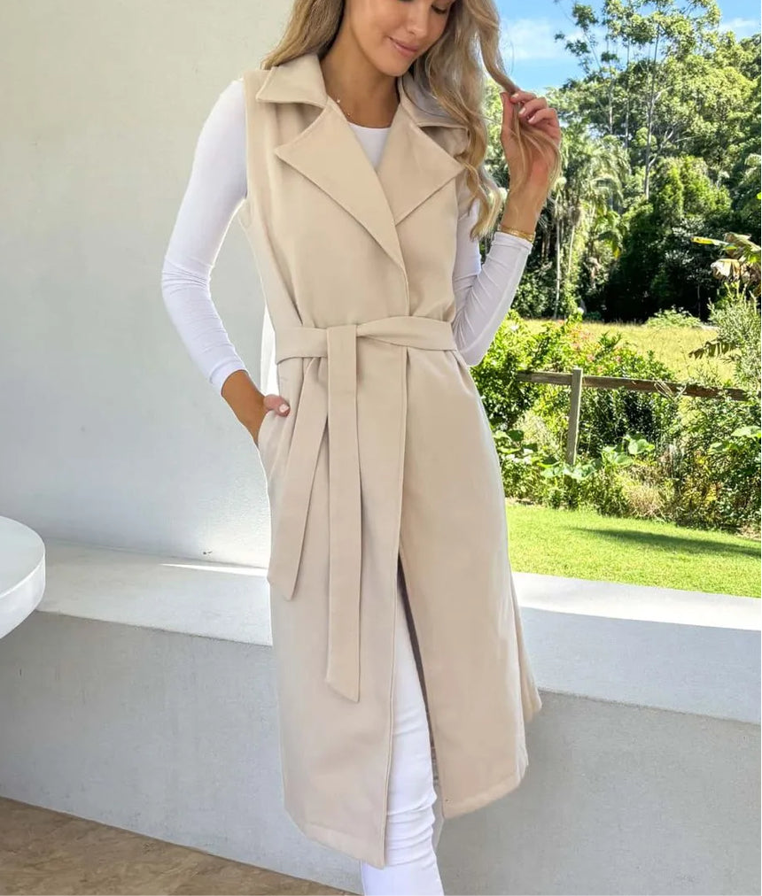 A woman wearing a beige sleeveless trench vest with a lapel collar and belted waist, paired with a white top and light-colored trousers.