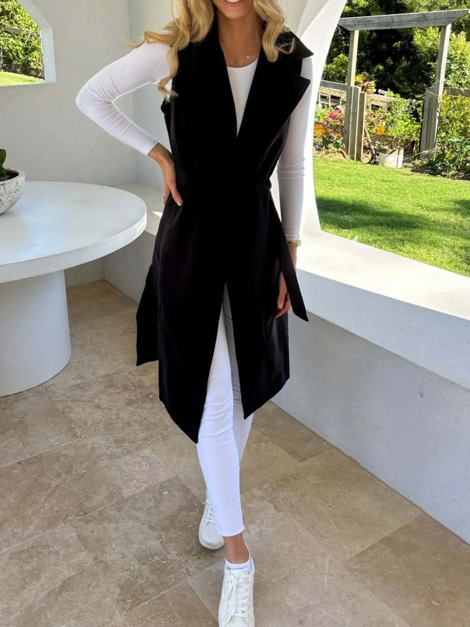 A woman with hand on waist, wearing a black sleeveless trench vest over a white top, with the vest's belt tied at the waist, standing by a window with a green view.