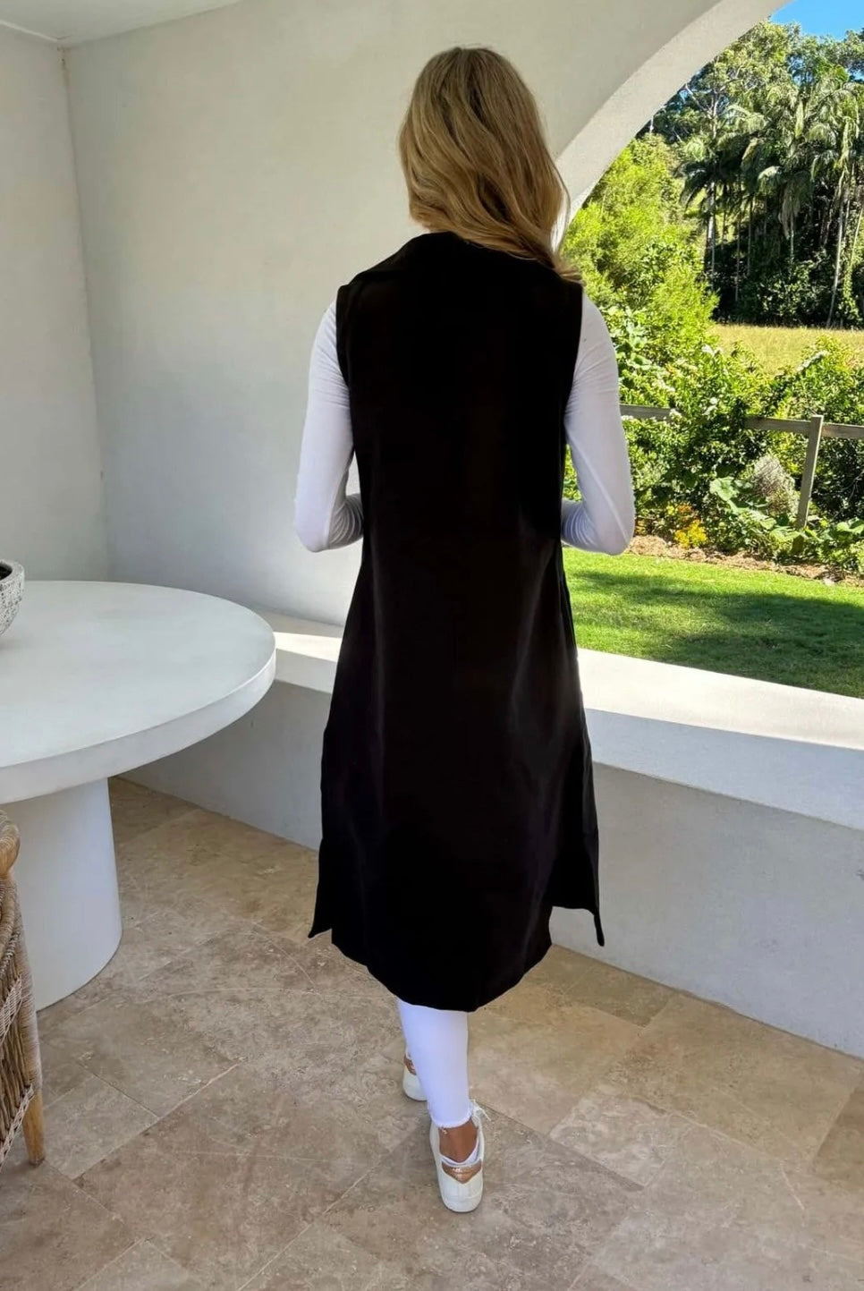 A back view of a woman wearing a black sleeveless trench vest over a white top, with the vest's belt tied at the waist, standing by a window with a green view.