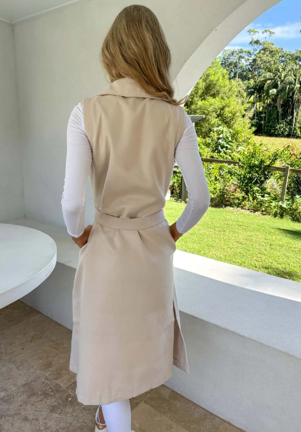 A woman wearing a beige sleeveless trench vest over a white top, with the vest's belt tied at the waist, standing by a window with a green view. Back View.