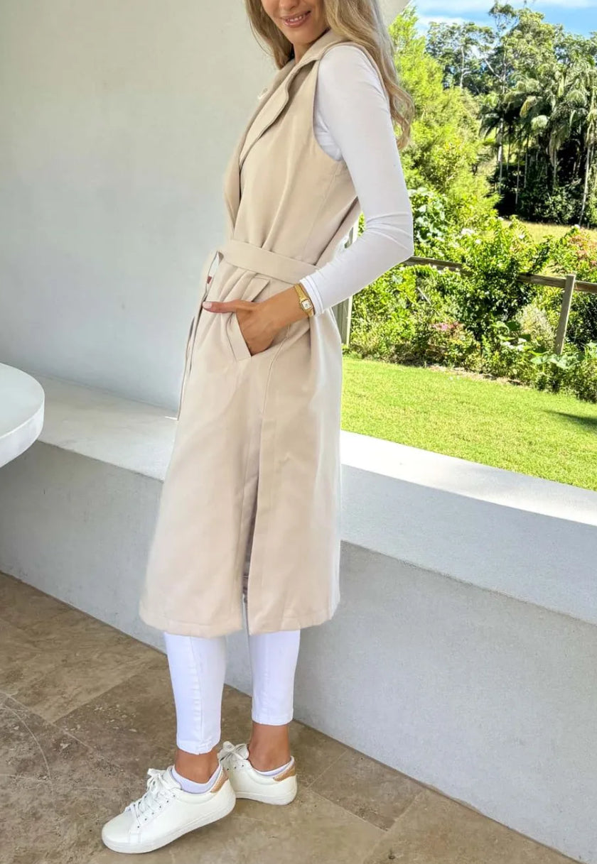 A woman wearing a beige sleeveless trench vest over a white top, with the vest's belt tied at the waist, standing by a window with a green view, side view.
