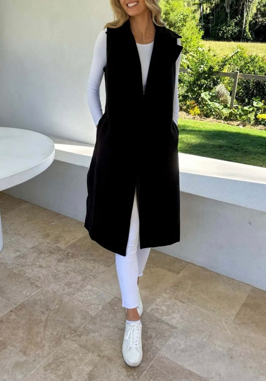 A woman with hands in pocket wearing a black sleeveless trench vest over a white top, with the vest's belt tied at the waist, standing by a window with a green view.