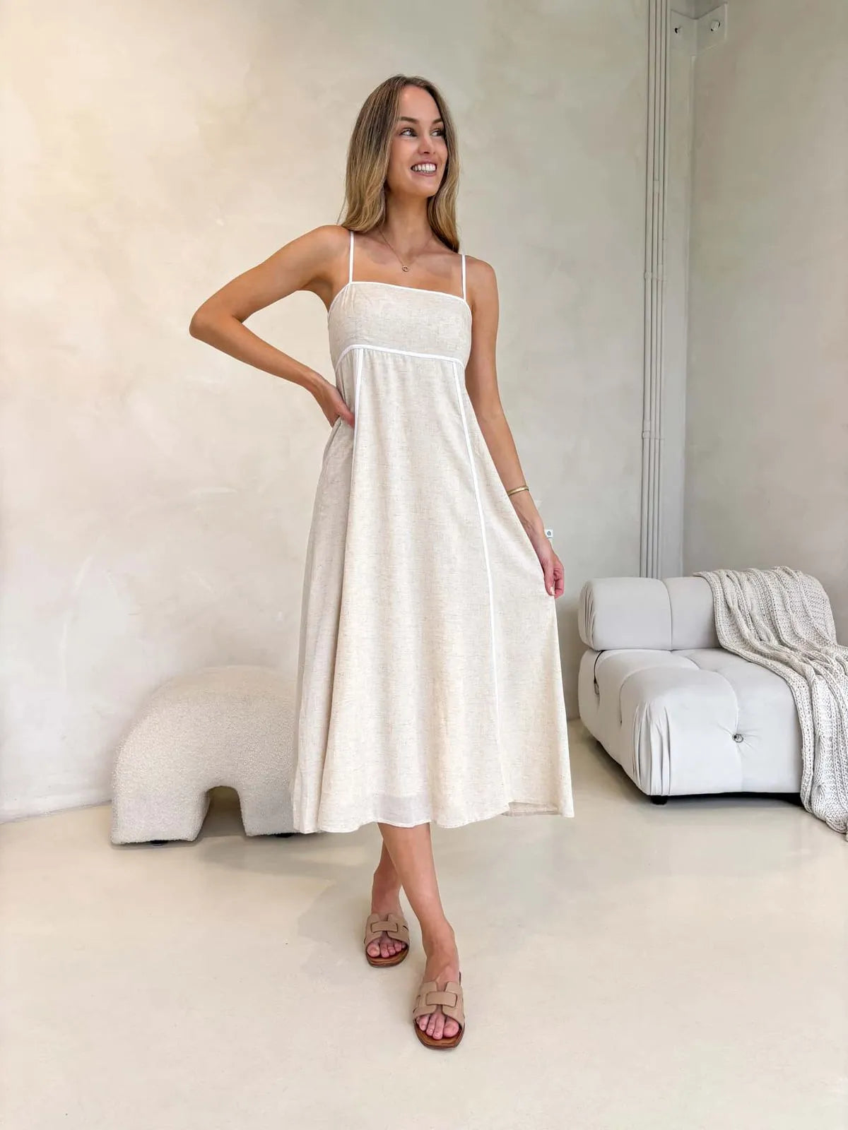 Model standing in a modern room wearing a natural-toned, flowy midi dress with white piping and spaghetti straps.