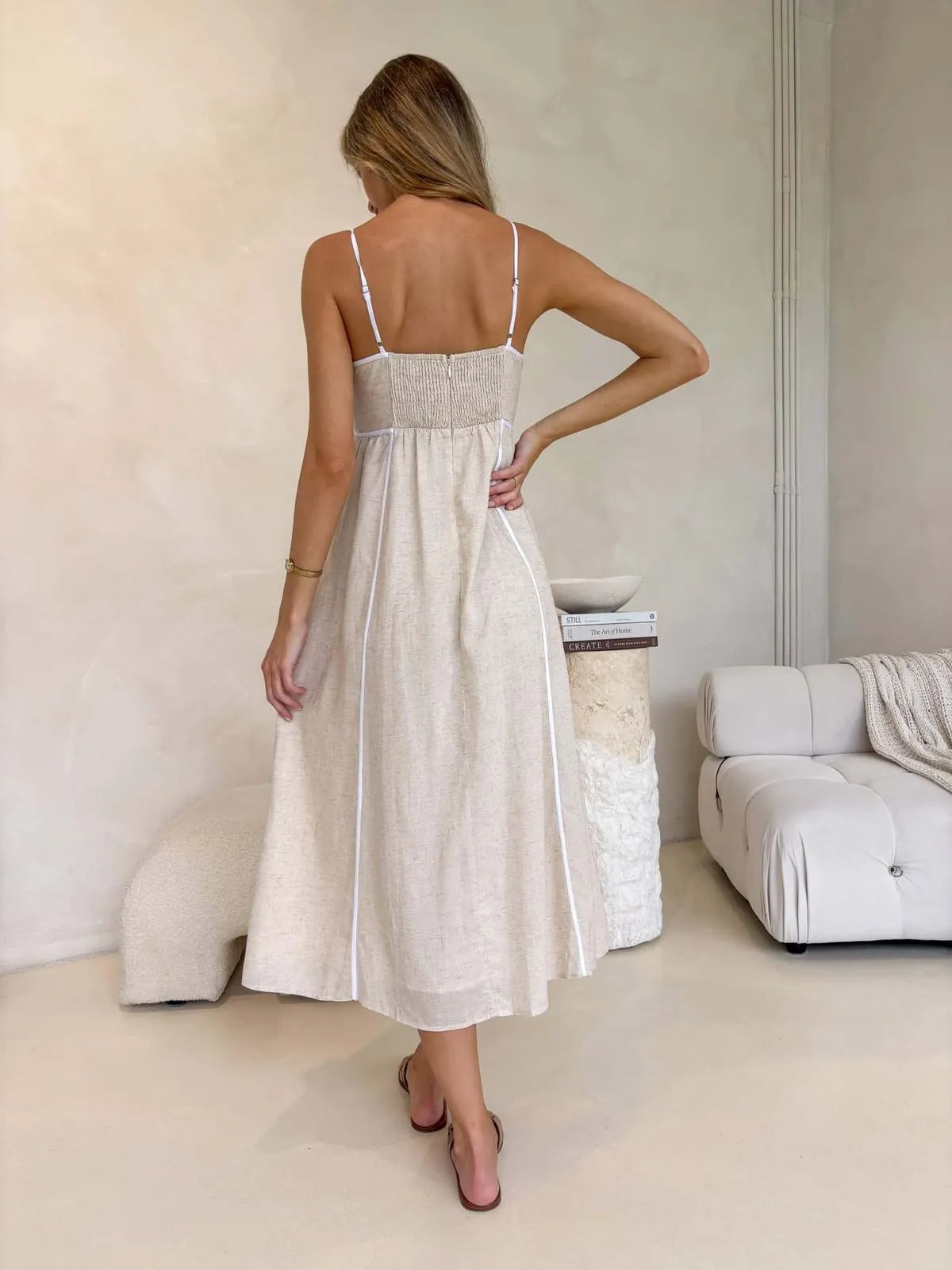 Back view of a owman in a neutral boho-style midi dress with shirred back and white trim, styled with flat sandals