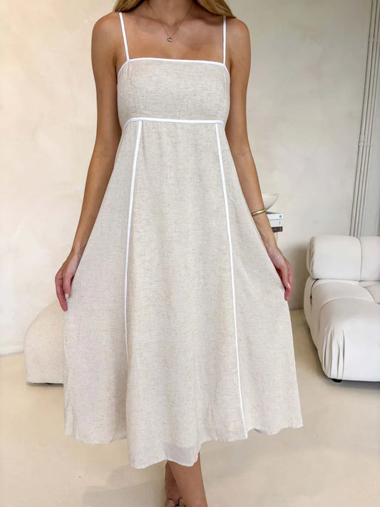 Woman wearing a natural linen look midi dress with white binding and thin straps, showcasing front detailing in a mininal indoor setting